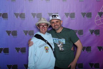 Two smiling people posing together in front of a purple backdrop with repeating white logos; one wears a light blue hoodie and cowboy hat, while the other wears a green t-shirt and a cap.