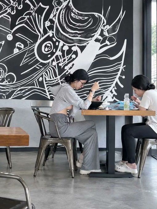 Two women sit at a café table, eating and using their phones, in front of a large black-and-white abstract mural. The accompanying post jokingly describes the pictured girlfriend “suddenly” punching her after a shopping trip, adding a playful narrative to the otherwise casual scene; no readable text is visible.