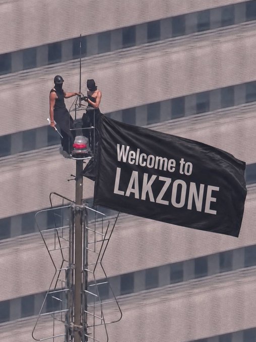 Two workers hoist a black "Welcome to LAKZONE" banner atop a pole against a high-rise building, marking the community's expansion as seven new members—including @0x_hamid, @Bigbbybenny, @GROOT__04, @Soliu08, @emptystdotcom, @k2sbhai, and @heis_Aiden—are welcomed to the platform.