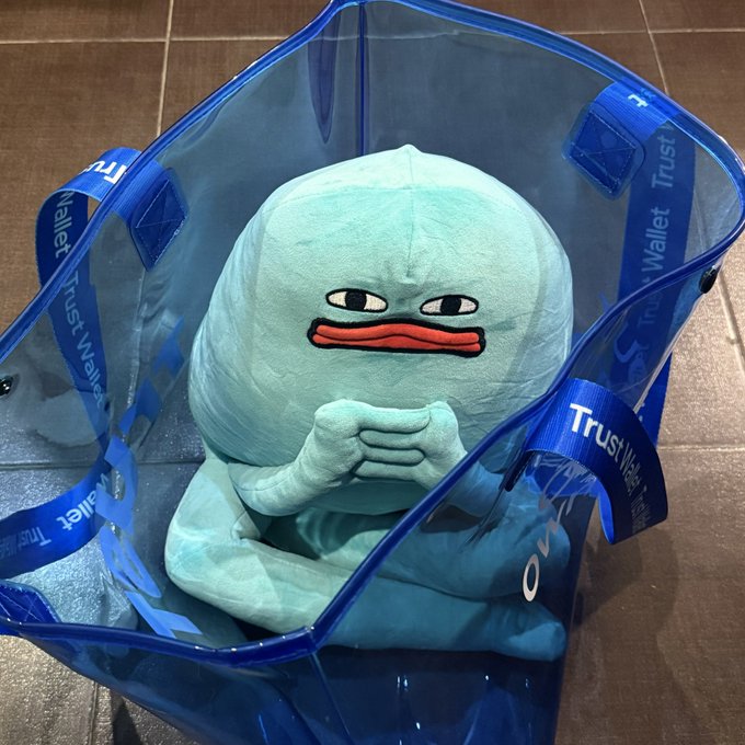 A light blue plush character with a skeptical expression sits inside a translucent blue tote bag with "Trust Wallet" branded straps.