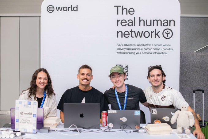 Four representatives of the "World" project pose behind a table displaying their laptops and a plush mascot at an ETHGlobal Lisbon event, standing in front of a banner promoting "The real human network" and its mission to verify unique humans online as AI advances.