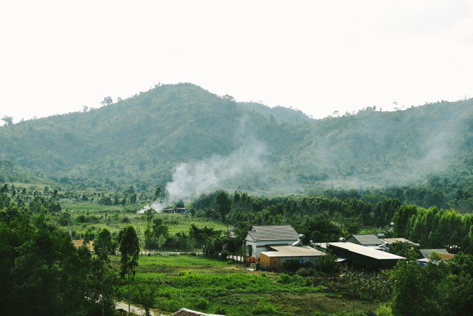 The image shows a beautiful mountain landscape with a small village nestled at its base, surrounded by lush green trees. The sky above appears hazy, adding a sense of tranquility to the scene. In the foreground, there are two houses visible, one larger and more prominent than the other. A person is standing in front of these houses, possibly admiring the view or attending to their property.