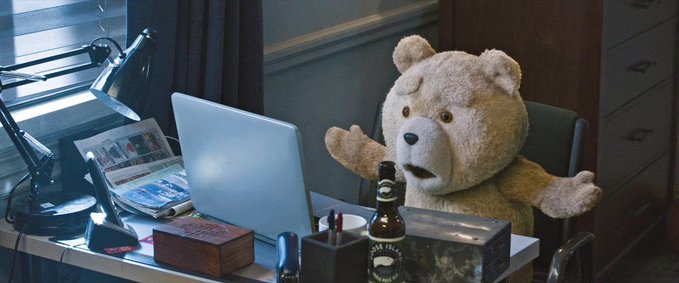Ted, the talking teddy bear from the Ted movies, sits at a desk with a laptop and a beer bottle, looking surprised.
