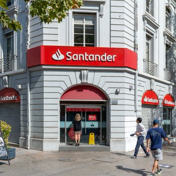 The image shows a Santander bank branch with its distinctive red logo prominently displayed on the facade, as pedestrians walk by on the sidewalk. This relates to the post about Santander, Spain's largest bank, holding $4 million in Bitcoin ETFs, highlighting the growing involvement of traditional banking institutions in cryptocurrency investments.
