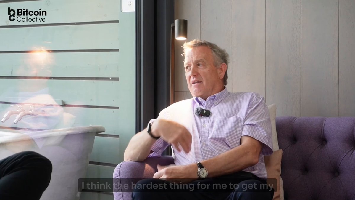 The image shows an older man sitting comfortably on a purple couch with his arm resting on his knee. He appears to be wearing a watch and has grey hair, suggesting that he might be in his sixties or seventies. The background of the image is blurred, but it seems like there are some windows present as well. There is also text visible in the image, which reads "DROPPING LATER TODAY Nicholas Dickinson has owned 13 hotels over 40 years. Nine months ago, he barely knew what Bitcoin was." This could be a part of a conversation or interview about Bitcoin and its impact on the real estate market.