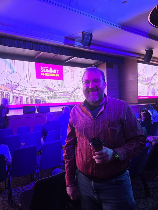 A bearded man in a plaid shirt stands in a conference venue holding a coffee cup, with a large screen behind him displaying 'THE SUMMIT' against a purple-lit backdrop.