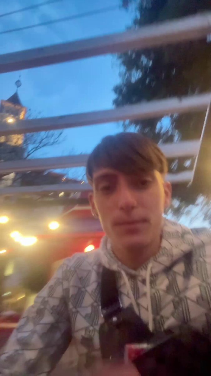 The image shows a young man wearing a white shirt that has black and gray designs, standing against a blue sky with white clouds. He appears to be enjoying himself while taking a selfie or looking at the camera. The photo is tagged with social media handles @LunarStrategy and @gyokeresethat are not immediately clear but seem to provide context for the content of the image. A black bag can also be seen on the right side, which might belong to the young man.