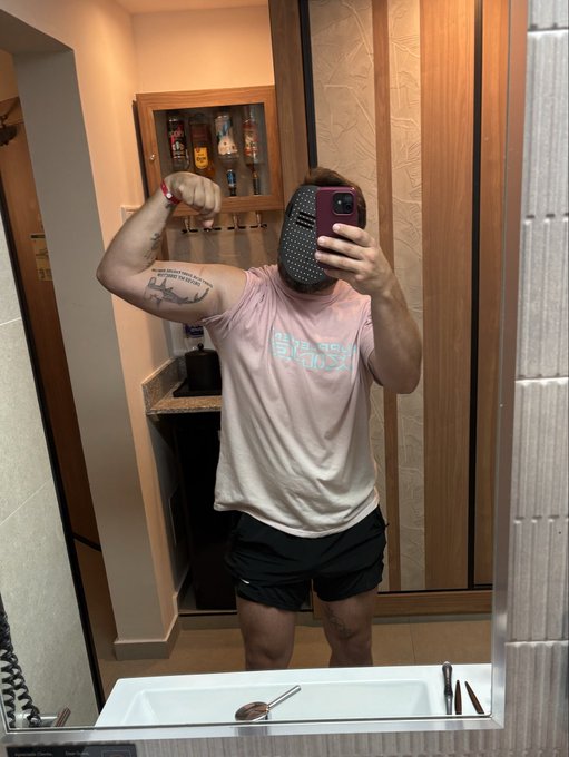 A person wearing a pink Stüssy t-shirt and black shorts takes a mirror selfie while flexing their arm, with their face obscured by a patterned object.