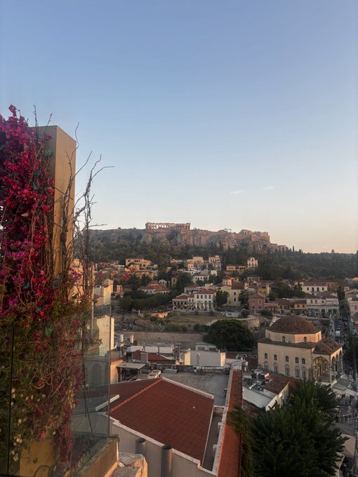 A scenic view from a balcony overlooking Athens, Greece, with the Acropolis visible on the hill in the distance and bougainvillea flowers in the foreground.