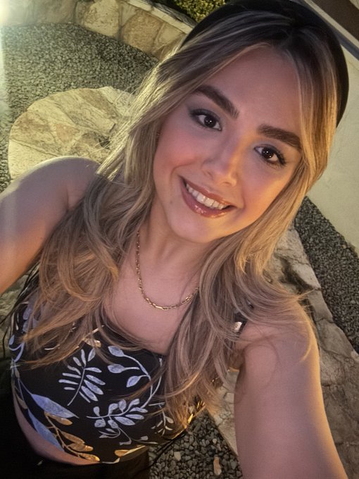 A selfie of a smiling young woman with long blonde hair wearing a black floral patterned top and a delicate gold necklace, with sunglasses pushed up on her head.