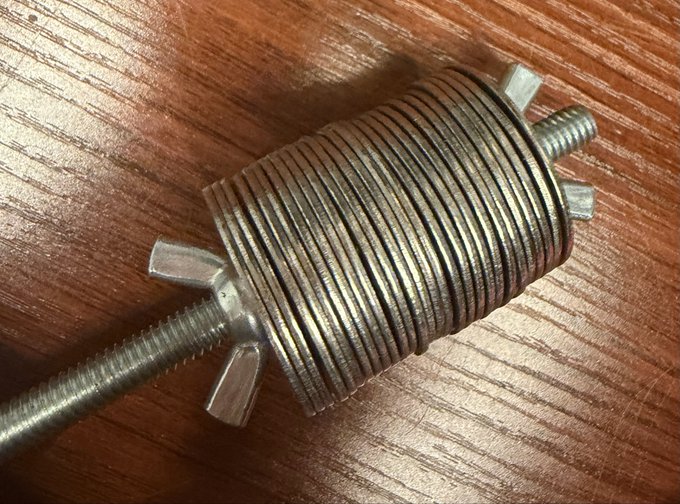 Close-up photograph of a threaded metal rod with a coiled spring wrapped around it and two wing nuts, lying on a wooden surface.