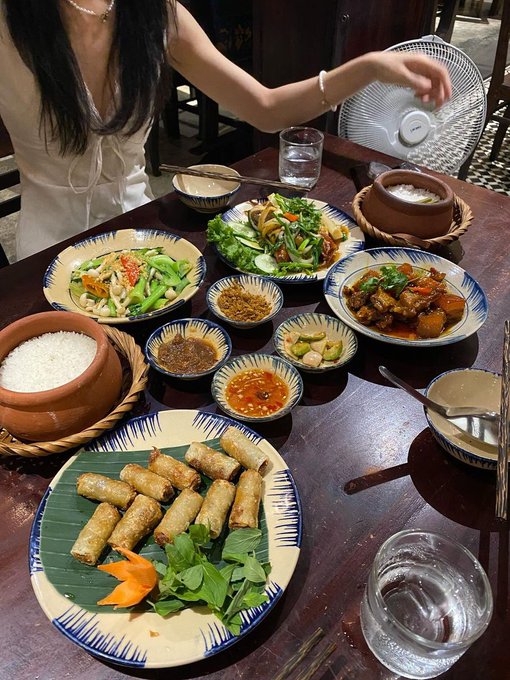 A table laden with various Vietnamese dishes including spring rolls on a banana leaf, rice in clay pots, and stir-fried vegetables, with a woman seated in the background.