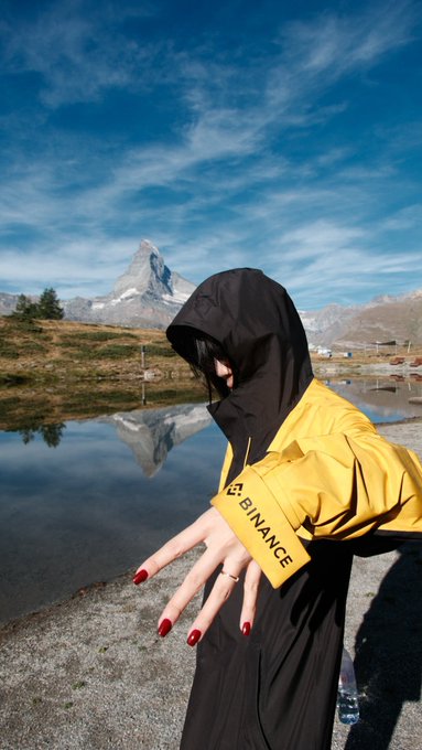 A person wearing a black hooded jacket holding a yellow Binance-branded umbrella, standing in front of a reflecting lake with the Matterhorn mountain visible in the background.