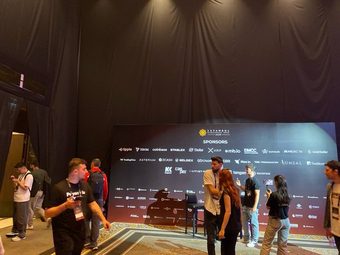 The image shows a group of people standing around a large screen displaying logos for various companies such as Bitcoin, Babylon, and TAVI. The people are engaged in conversation with each other while some are holding drinks or using their cell phones. The event appears to be an informal gathering where attendees can discuss these topics and socialize.