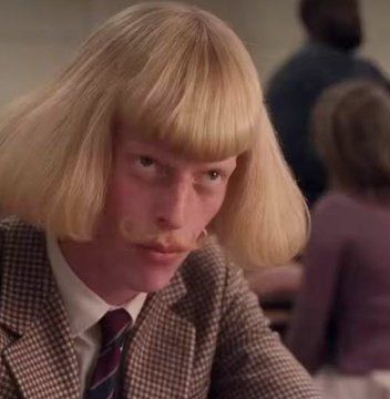 A man with a blonde bowl haircut and matching mustache stares forward with an intense expression, wearing a tweed jacket and striped tie.