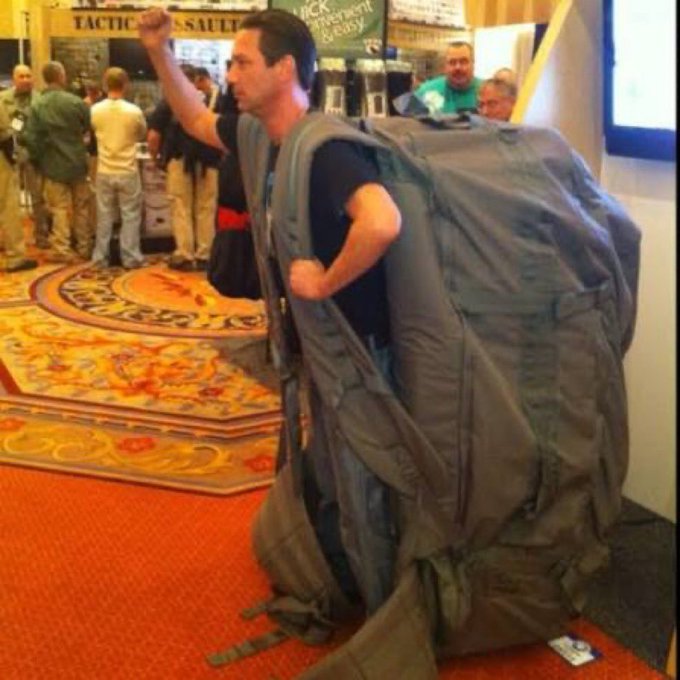 A man at what appears to be a tactical gear trade show (with "TACTICAL" visible on a banner behind him) is humorously wearing an oversized olive-drab backpack that's nearly as tall as he is, pumping his fist in the air as if ready to charge forward. The image visually embodies the post's message about preparing for an upcoming bull market—suggesting the man (and investors) are fully geared up and eager to move forward with confidence.