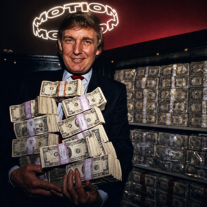 A man in a suit smiling while holding a large stack of banded $50 bills, standing in front of a wall of money bricks with a neon 'Motion' sign overhead.