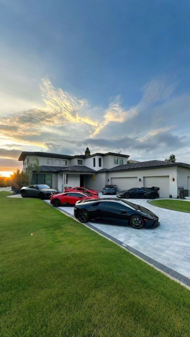 A modern luxury home with multiple sports cars, including a black Lamborghini and red Porsche, parked in the driveway at sunset.
