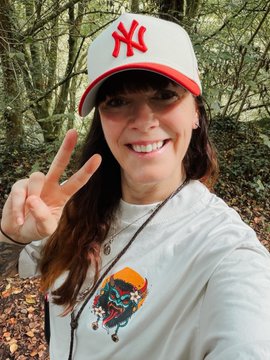 A smiling woman wearing a New York Yankees cap and a white t-shirt with a colorful graphic makes a peace sign outdoors.