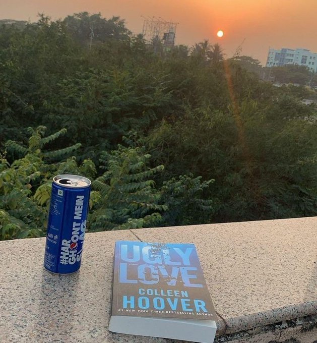 The image features a book titled "Ugly Love" by Colleen Hoover, with a blue can of soda placed next to it on a ledge or balcony. The scene suggests that someone has been reading the book while enjoying their drink outside during sunset. The presence of the book and the soda together indicates that they might be taking a break from work or other activities to relax and enjoy some leisure time outdoors, possibly discussing the content of the book with each other or simply appreciating the beauty of the sunset.