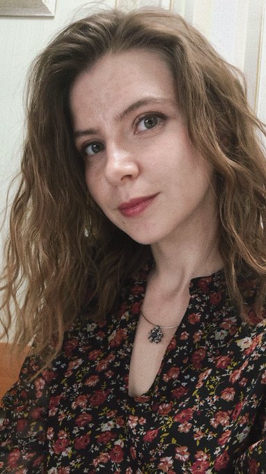 A close-up portrait of a young woman with long, wavy light brown hair, wearing a dark floral-patterned top and a small pendant necklace.