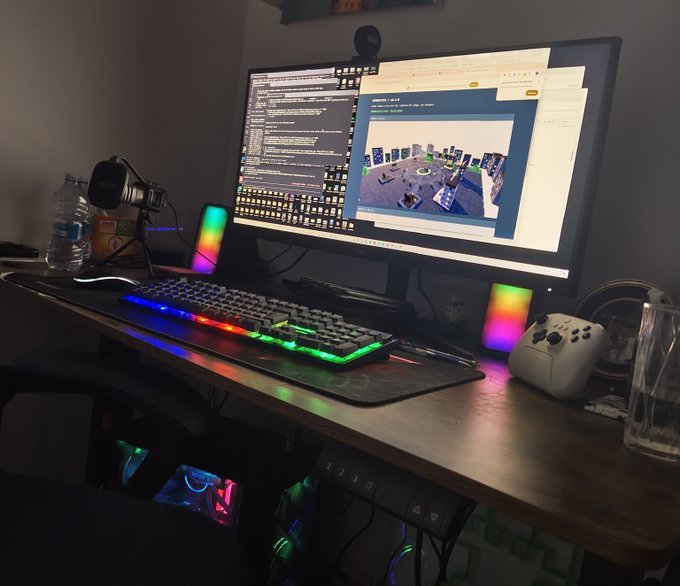 A dimly lit gaming/computer setup featuring a monitor displaying code and a 3D environment, a rainbow-backlit mechanical keyboard, RGB speakers, and a white controller on a wooden desk.