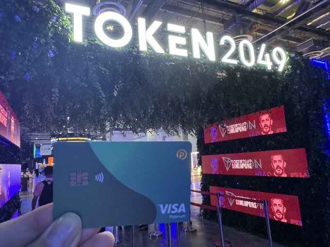 A hand holds a teal Visa Platinum card in front of an illuminated 'TOKEN2049' sign at a conference, with red banners reading 'TOKEN2049 SINGAPORE' featuring a man's face in the background.