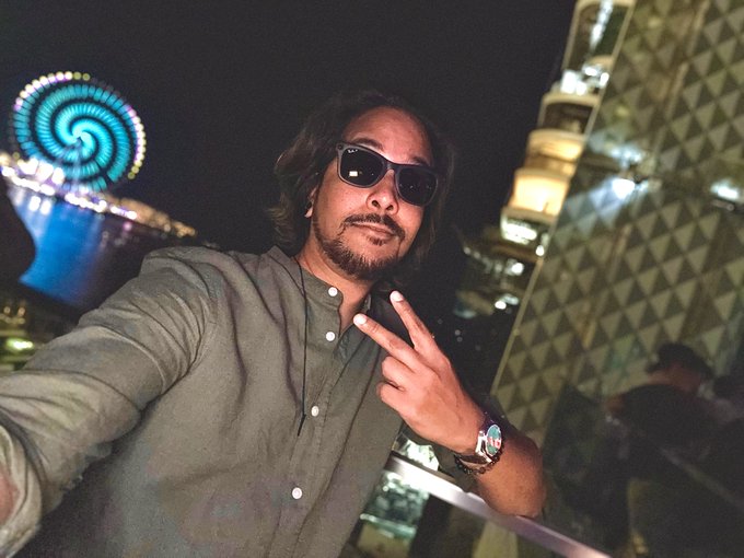 A man with long hair, sunglasses, and a gray shirt makes a peace sign while taking a selfie at night, with a brightly lit Ferris wheel and modern architectural structure visible in the background—capturing a stylish "good night" moment.