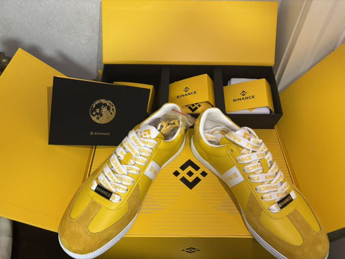 A pair of yellow Binance-branded sneakers with white laces, displayed on top of and inside a yellow Binance shoebox.