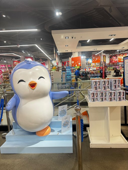 A large inflatable penguin mascot with a cheerful expression stands on a blue platform inside a brightly lit retail store, surrounded by stanchions and displays of merchandise including boxed products with face imagery. The store appears to be a souvenir or character-themed shop with colorful displays visible in the background.