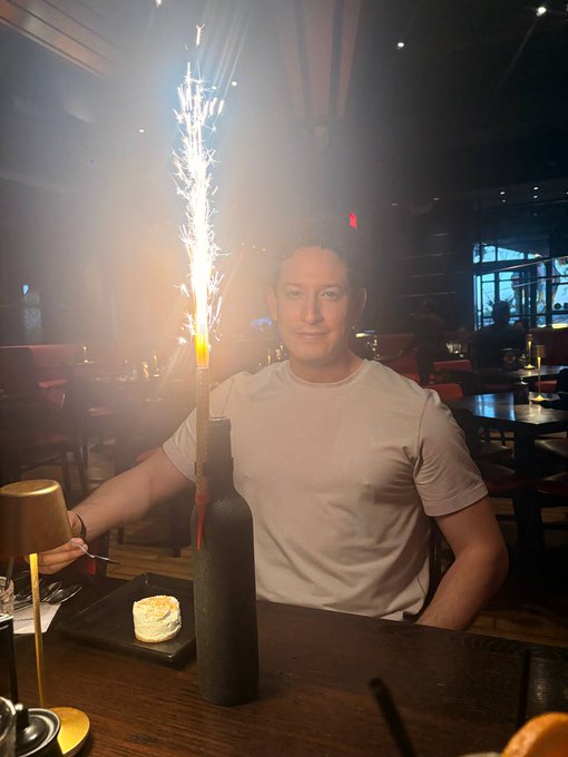The image features a man sitting at a table with a cake placed next to him. The cake has white frosting, and there are candles on top of it. A bottle containing sparklers can be seen nearby as well. In the background, there is another person seated at a different table.