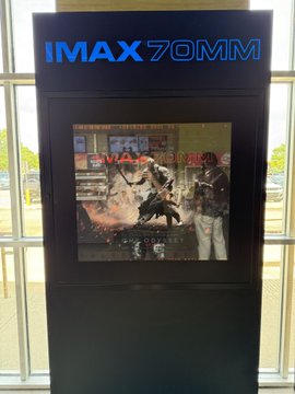 An IMAX 70MM display board shows a movie poster featuring "MA" (likely "Mad Max") and reads "NOW SHOWING IN IMAX 70MM," fitting the poster's caption of being seated and ready for the immersive IMAX experience.