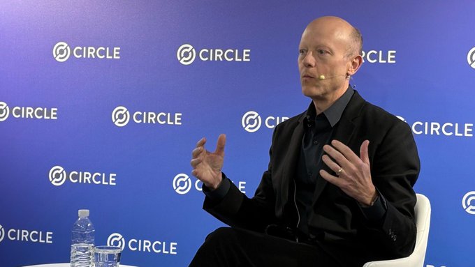 A bald man in a black suit gestures while speaking on stage at a Circle (cryptocurrency company) event, with the Circle logo repeated on the blue backdrop behind him.