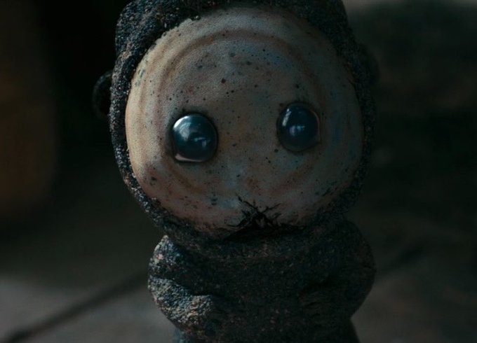 A close-up of a creepy, weathered doll-like character with large blue eyes, wearing a dark hooded garment, from what appears to be a horror film or game.