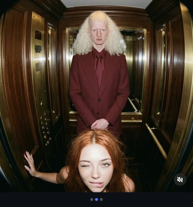 A person with pale skin and long curly blonde hair wearing a maroon suit stands in a wood-paneled elevator behind another person with red hair who is winking at the camera.