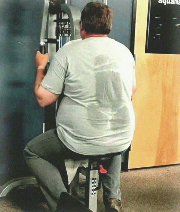 A man is shown from behind, sitting on exercise equipment in a gym, with a noticeably large sweat stain visible on the back of his gray t-shirt. Part of the word 'aquamar' is visible on a window or sign in the upper right corner.
