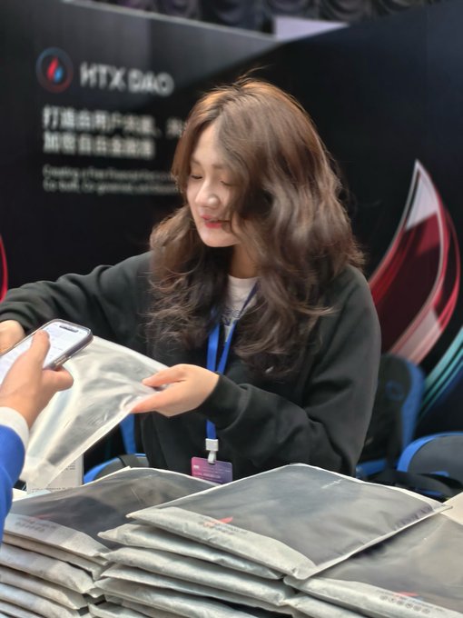 A young woman with long wavy hair hands out silver promotional packages at an HTX DAO booth, with the company's logo and bilingual Chinese/English branding visible on the backdrop. The scene visually ties to the post's theme of HTX DAO actively engaging its community and working to create tangible value for platform token holders during a challenging market period.