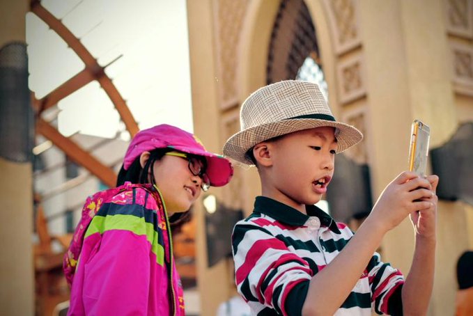 Two children wearing sun hats look at a smartphone together, with the boy in a straw fedora holding the device and the girl in a pink jacket beside him.