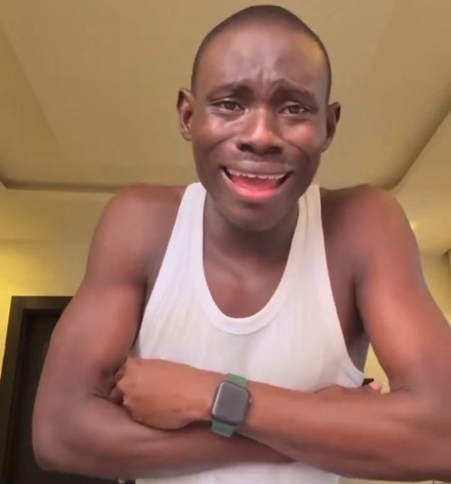 A close-up photo of a young man wearing a white tank top and a smartwatch, with a surprised or emotional facial expression, indoors.