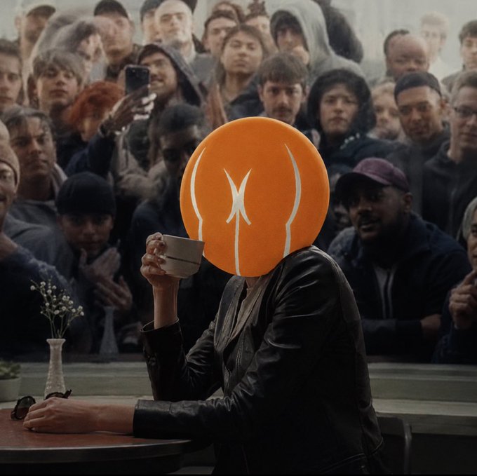The image features a person wearing an orange circle headdress, sitting at a table with their hands on their lap while holding a cup of coffee or tea. The person's face is partially obscured by the headdress, making them appear as if they are part of the crowd in front of them. The background shows several people standing and looking towards the camera, creating an atmosphere of a public gathering or event.