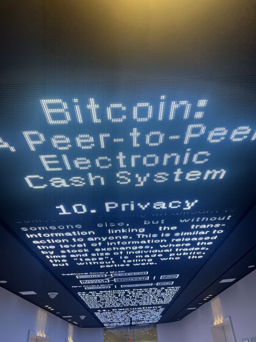 An illuminated display board showing the text 'Bitcoin: A Peer-to-Peer Electronic Cash System' alongside an excerpt from its '10. Privacy' section.