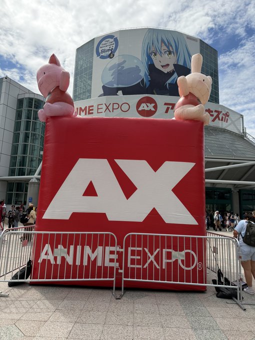 An outdoor display at Anime Expo featuring a large red 'AX' sign with 'ANIME EXPO' text, flanked by inflatable characters, with a billboard for 'That Time I Got Reincarnated as a Slime' visible behind it.
