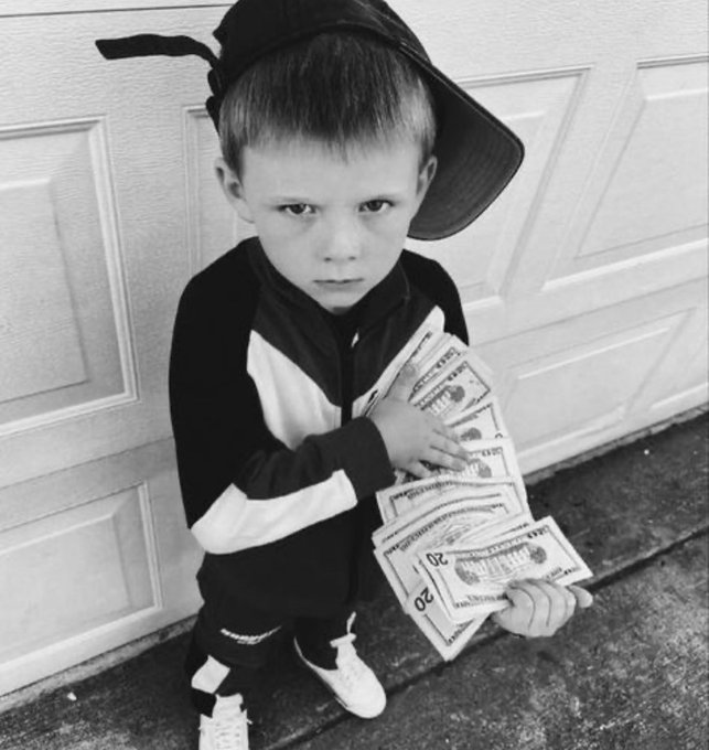 A black-and-white photo of a young boy wearing a backwards cap and tracksuit, holding a fan of twenty-dollar bills.