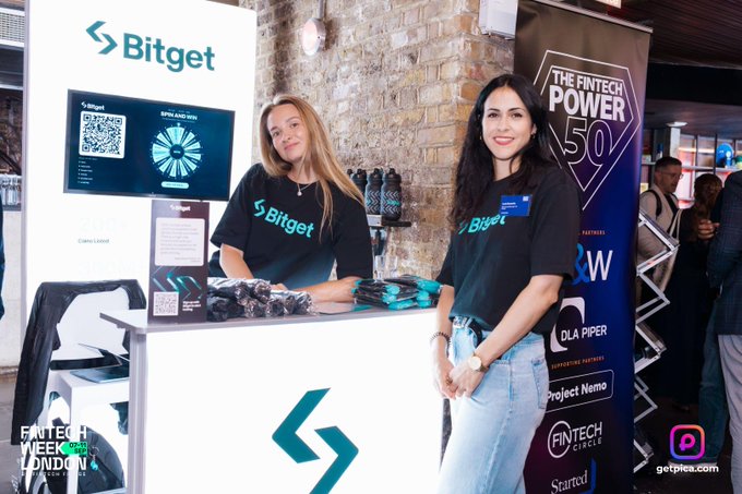 Two women wearing dark green "Bitget" t-shirts stand behind a promotional counter at an event. Banners for "Bitget" and "THE FINTECH POWER 50" are visible, with text indicating "FINTECH WEEK LONDON" and "getplex.com".