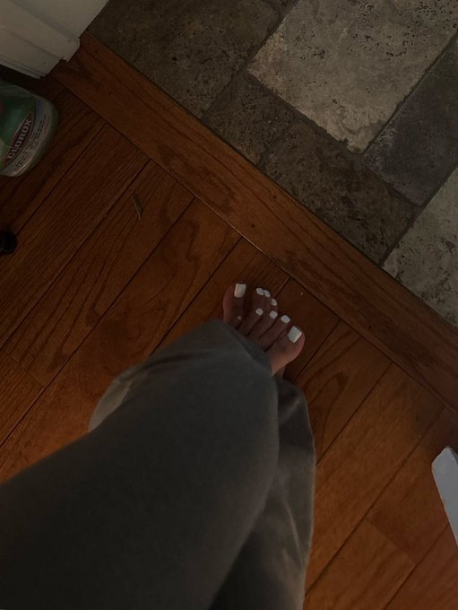 A first-person view shows a person with a fresh white pedicure standing on a hardwood floor next to a tiled threshold, with a bottle of Clorox visible in the corner, perfectly showcasing their newly painted toes.