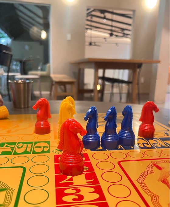 Colorful plastic horse game pieces (red, yellow, and blue knights) are arranged on a yellow Ludo board in a room with a dining table visible in the background.