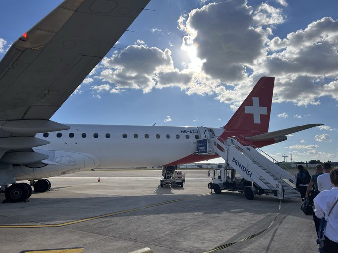 Passengers boarding a Brindisi airplane on the tarmac under a partly cloudy sky with the sun shining through clouds.