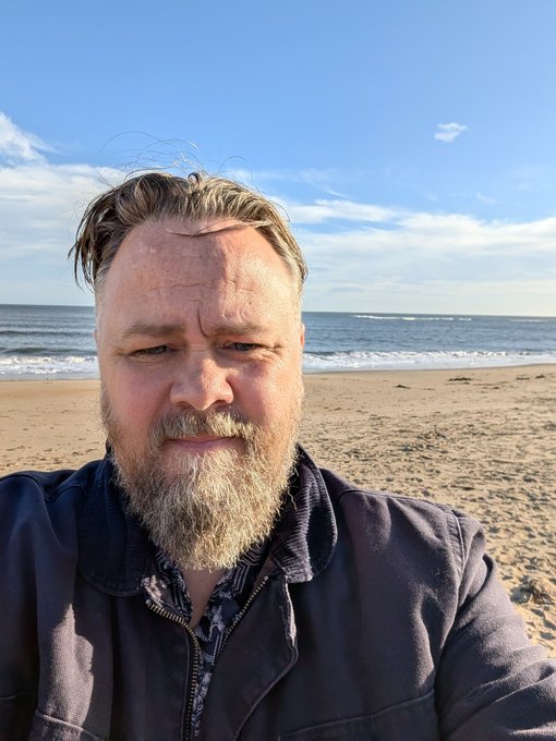 A selfie of a bearded older man in a dark jacket standing on a sandy beach with the ocean and a partly cloudy sky behind him.