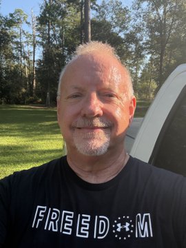 A smiling man wearing a black "FREEDOM" t-shirt takes a selfie outdoors with a grassy area, trees, and a white vehicle in the background, fitting the caption "More miles for the" to suggest a road trip or travel vibe.