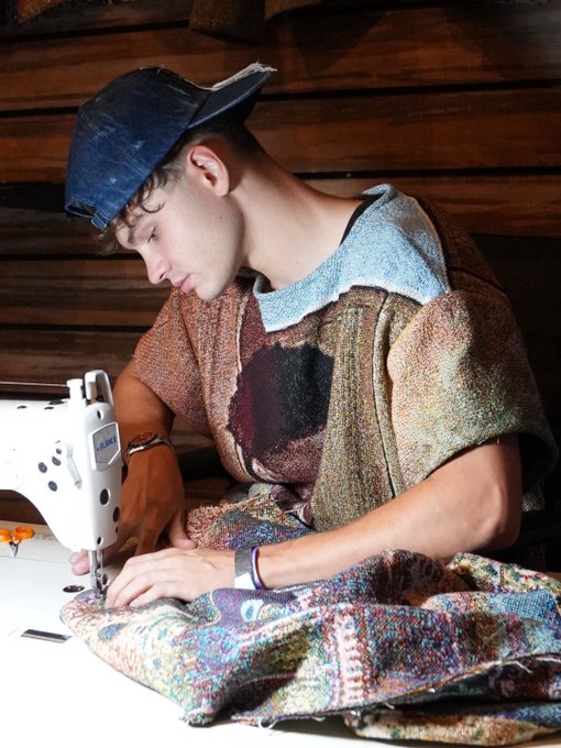 A young man wearing a blue denim cap and a patchwork sweater sews fabric on a white sewing machine.